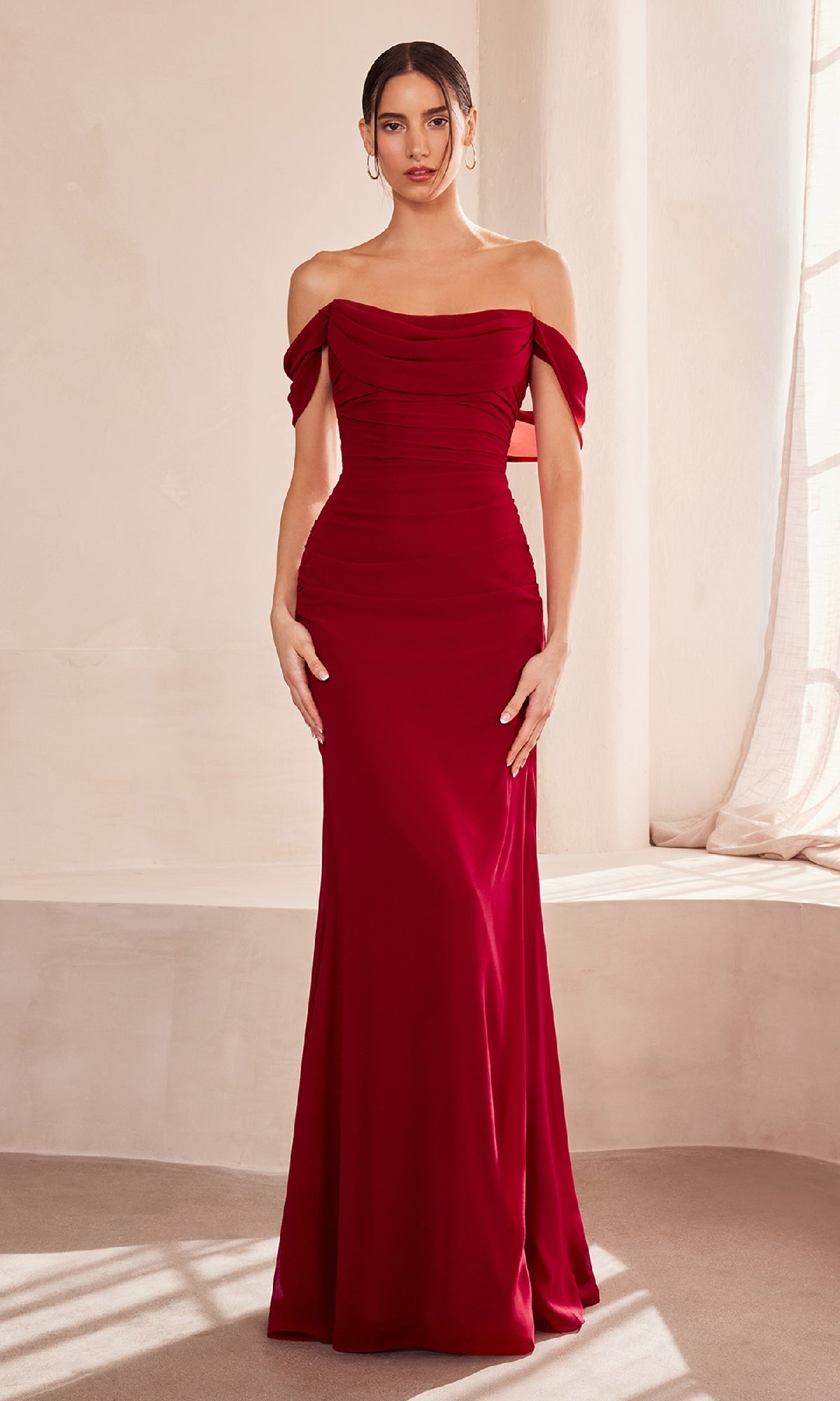 Indoors, a woman wears the Off-Shoulder Classic Prom Gown: Amoris T705, a fitted, floor-length red dress with ruched detailing—elegant and affordable, perfect for special occasions.